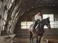Une cavalière en tenue d'équitation monte un cheval brun dans un manège couvert. Le cheval est au trot, prêt pour une séance de demi pension cheval.