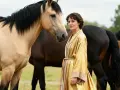 Une femme en robe dorée se tient près d'un cheval isabelle et d'autres chevaux dans un pré.