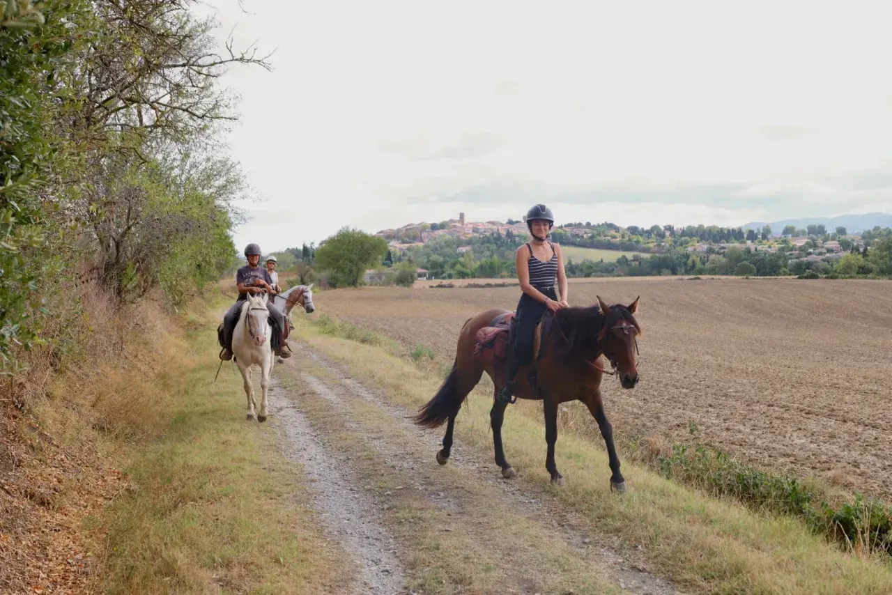 Trois cavaliers profitent d'une promenade à cheval sur un chemin de terre, avec un village en arrière-plan.