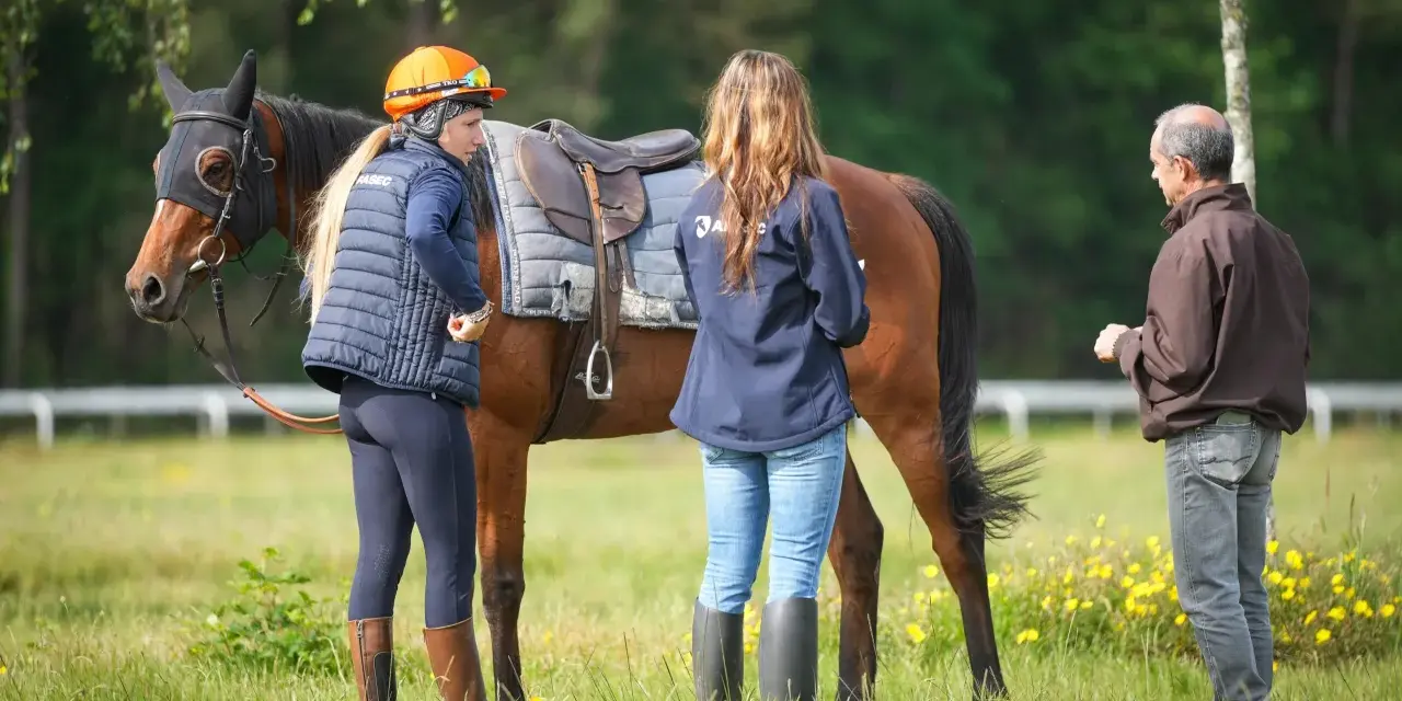 Une femme en tenue d'équitation discute avec un homme près d'un cheval sellé, lors d'une formation cheval pour adulte en extérieur.