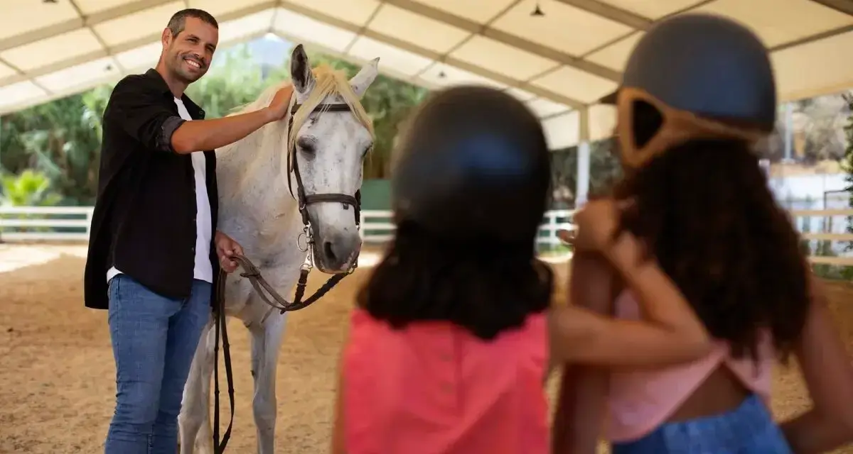 Un instructeur d'équitation souriant caresse un cheval blanc. Deux enfants, casqués, regardent.