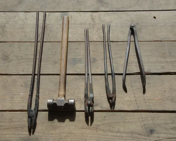 Ensemble d'outils de maréchal-ferrant : pinces, marteau et autres instruments sur fond de bois.