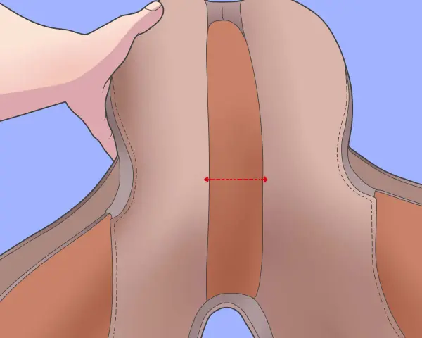 Une main tient une selle de cheval. Une flèche rouge indique comment mesurer la largeur de l'ouverture de la selle, une étape clé pour savoir comment mesurer une selle de cheval.