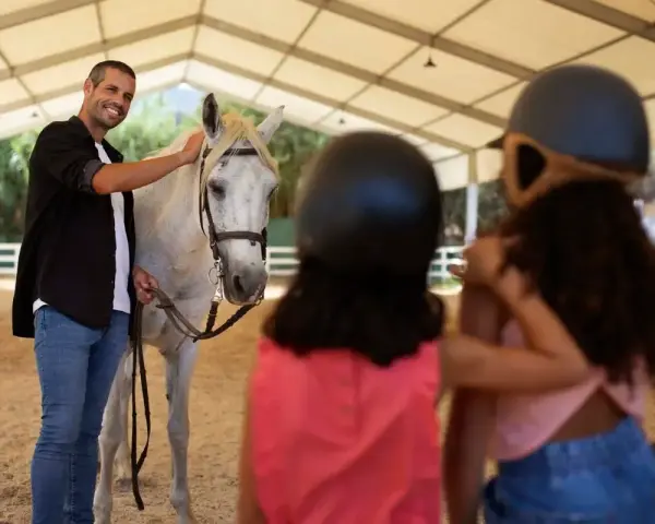 Un instructeur d'équitation souriant caresse un cheval blanc. Deux enfants, casqués, regardent.