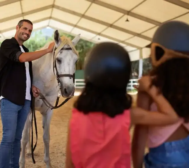 Un instructeur d'équitation souriant caresse un cheval blanc. Deux enfants, casqués, regardent.