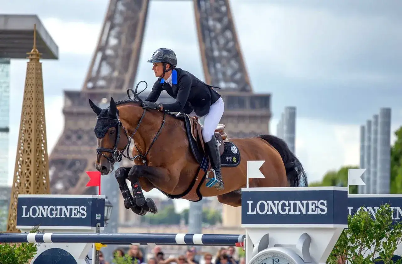 Cavalier et cheval brun sautent un obstacle devant la Tour Eiffel. La flora de mariposa s'épanouit dans ce décor parisien.