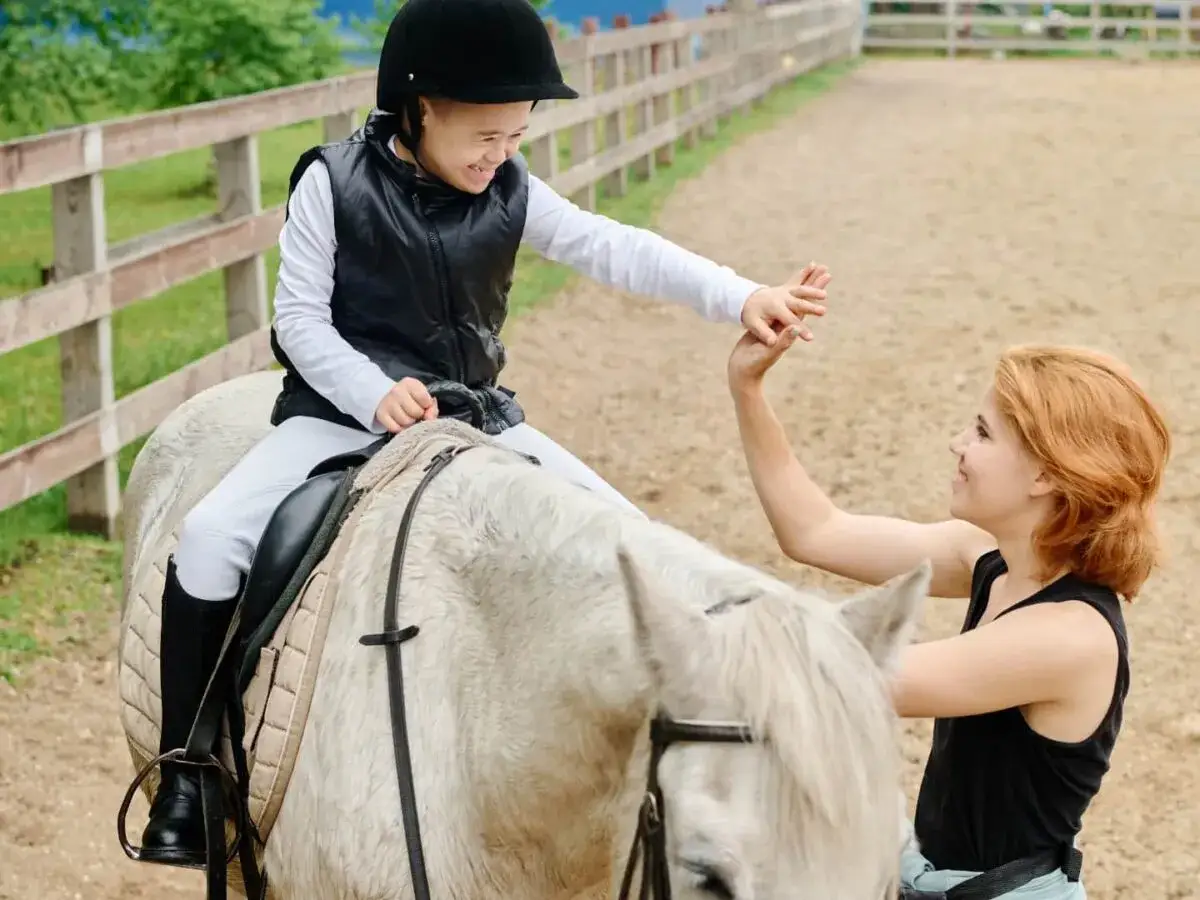 Une jeune fille souriante, casquée, monte un cheval blanc. Son instructeur d'équitation lui tend la main, l'encourageant.
