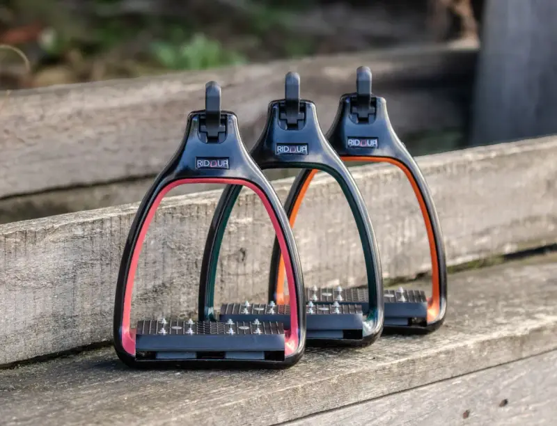 Trois étriers Rid'Up colorés (rose, vert, orange) sur un banc en bois. Avis positifs pour ces étriers.