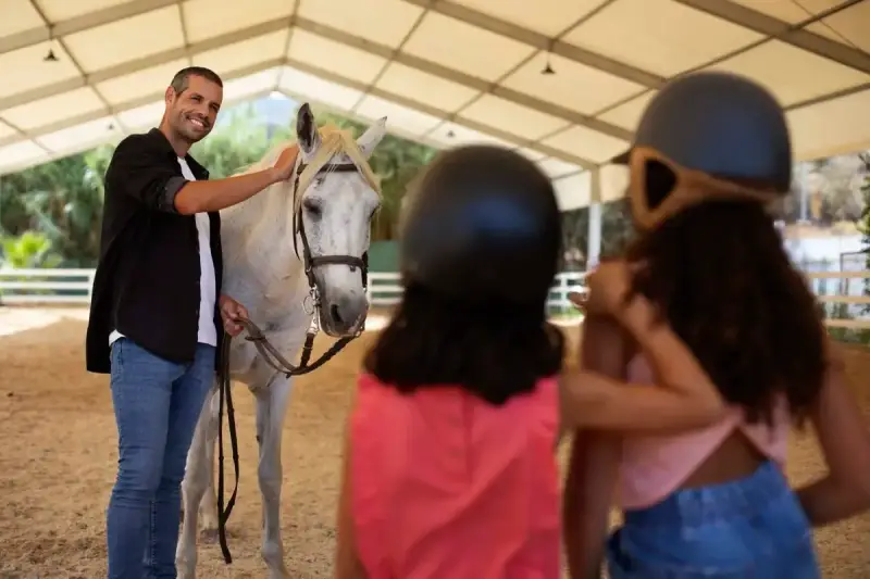 Un instructeur d'équitation souriant caresse un cheval blanc. Deux enfants, casqués, regardent.